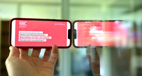 Ein Handy wird schräg gegen ein Spiegel gehalten. Auf dem Display sieht man die Schrift "Fakes und Manipulation: Was sollten wir tun, um unsere  Demokratie zu schützen?"