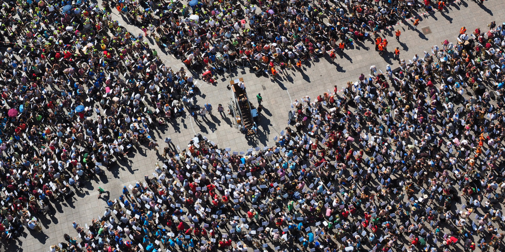 A huge crowd has assembled around a tall lectern on a town square. The crowd is shown from bird's eye view.