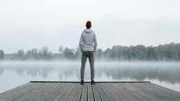 Ein Mann steht auf einem Steg an einem See, dessen Wasseroberfläche leicht im Nebel liegt. Der Mann ist von hinten zu sehen, wie er über den See hinweg in die Ferne blickt.