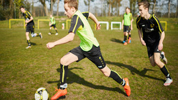 Ein junger Fußballer jagt auf dem Platz dem Ball hinterher, verfolgt von einem Gegenspieler. Im Hintergrund laufen weitere Spieler.