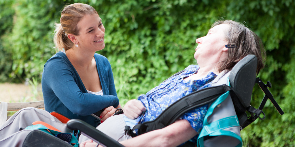 Ein Pflegebedürtige im Rollstuhl und ihre Pflegerin sitzen im Grünen und schauen sich an.