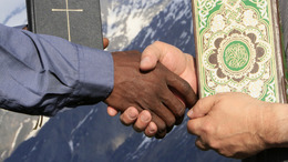 Ausschnitt zweier Menschen, die sich die Hände schütteln. In der zweiten Hand halten sie eine Bibel und einen Koran.