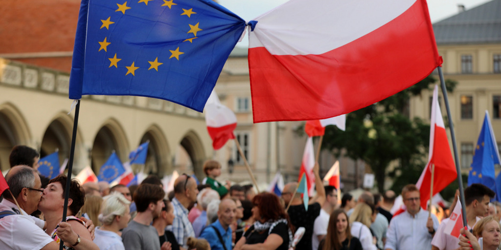 Eine große Menschenmenge mit Fahnen in der Hand, auf einem Platz, darüber befinden sich die Europaflagge sowie die polnische Flagge. In Hintergrund ein historisches Gebäude