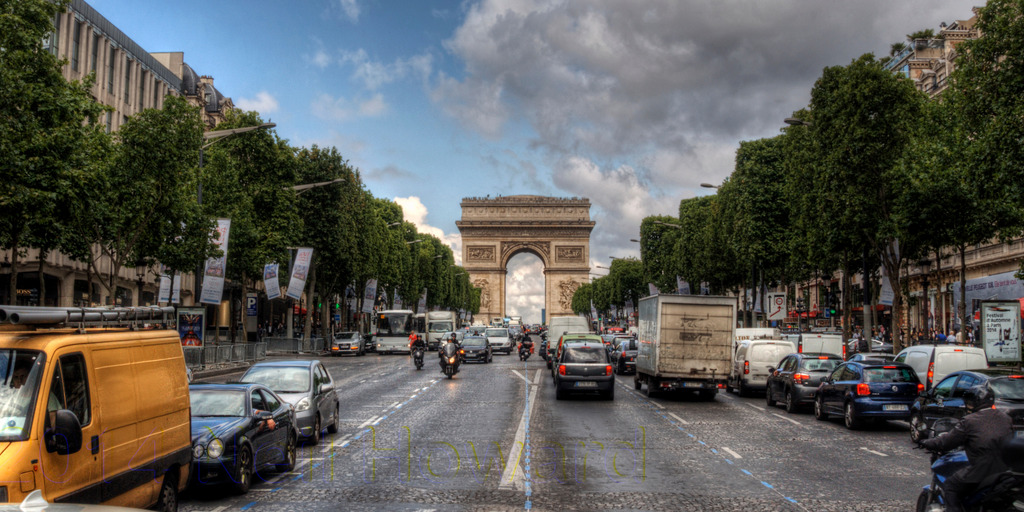 Blick auf den Champs Elysees in Paris. Im Hintergrund der Triumphbogen. Davor die Straße, befahren von Autos, LKWs und Mopeds.