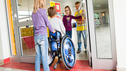 Ein Mädchen schiebt einen Jungen, der im Rollstuhl sitzt in Richtung einer Eingangstür, die von zwei anderen Kindern aufgehalten wird.