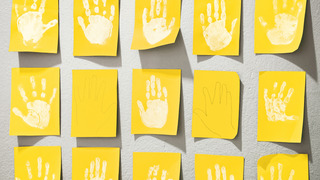 Weißfarbige Abdrücke von Kinderhänden auf gelben Pappkarten hängen an einer Wand