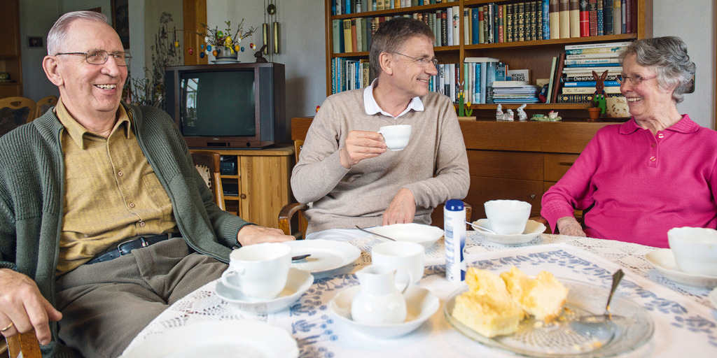 Ein älteres Ehepaar sitzt in seinem Wohnzimmer mit dem Hausarzt bei Kaffee und Kuchen.