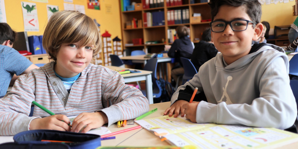 Zwei Schüler der Friedenauer Gemeinschaftsschule, Berlin, einem der Gewinner des Jakob Muth-Preises 2019, sitzen mit Stiften in der Hand vor ihren aufgeschlagenen Heften im Klassenraum und lächeln in die Kamera.