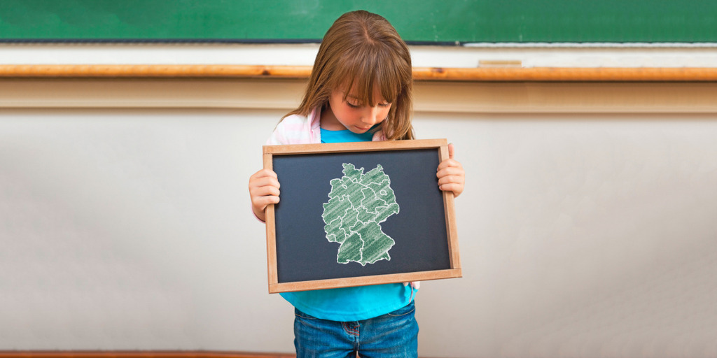 Ein Mädchen blickt auf eine kleine Tafel, die es in seinen Händen hält und auf der eine Deutschlandkarte mit den Umrissen der 16 Bundesländer aufgezeichnet ist. Im Hintergrund eine große Schultafel.