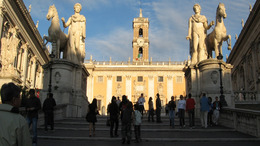Blick von den Treppen, die hinauf zum Kapitolshügel in Rom führen, auf den Senatorenpalast. Links und rechts säumen zwei große Statuen mit Pferden den Weg. Auf der Treppe laufen Menschen.