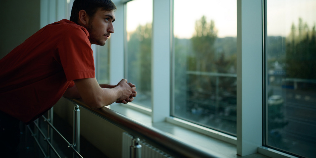 Ein junger Mann lehnt an einem Fensterbrett und schaut nachdenklich aus dem Fenster.