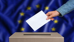 Eine Person wirft einen Stimmzettel in eine Wahlurne, hinter der eine Europafahne aufgehängt ist.