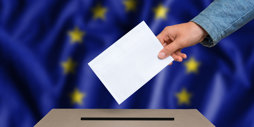 Eine Person wirft einen Stimmzettel in eine Wahlurne, hinter der eine Europafahne aufgehängt ist.