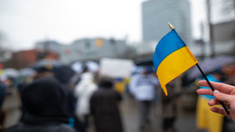 In a big city whose buildings can be seen in the background, a group of people protests against the war in Ukraine. A person in the foreground is holding a small Ukrainian flag into the camera.
