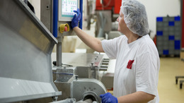 Eine Mitarbeiterin einer Lebensmittelfabrik trägt Arbeitskleidung, Handschuhe und ein Haarnetz und steht an einer Maschine, die sie über einen daran befestigten Touchscreen bedient.