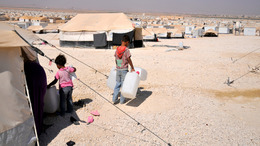 [Translate to English:] Blick auf das jordanische Flüchtlingslager Zaatari. Zu sehen sind hunderte befestigte Unterkunfszelte, die bis zum Horizont reichen. Im Vordergrund stehen zwei Kinder mit Wasserkanistern, die der Kamera den Rücken zugedreht haben