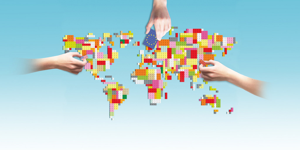 Weltkarte aus Legosteinen dargestellt. Drei Hände mit jeweils einen Legostein (Legostein mit der EU Flagge, China Flagge und USA Flagge) sind an der Ost-, West- und Nordseite der Weltkarte zu sehen.