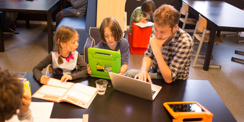 Ein Lehrer und zwei Schüler sitzen in einer Grundschule an einem Tisch. Der Lehrer und einer der Schüler lesen auf ihren Laptops, die zweite Schülerin hat ein Buch vor sich liegen und blickt auf den Laptop ihres Mitschülers hinüber.