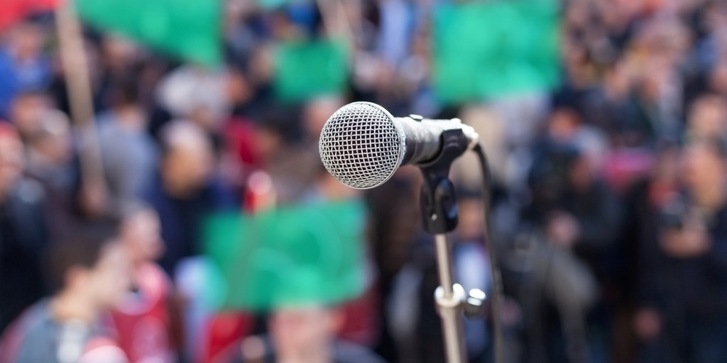 A microphone can be seen in front of a crowd which is holding up green placards.