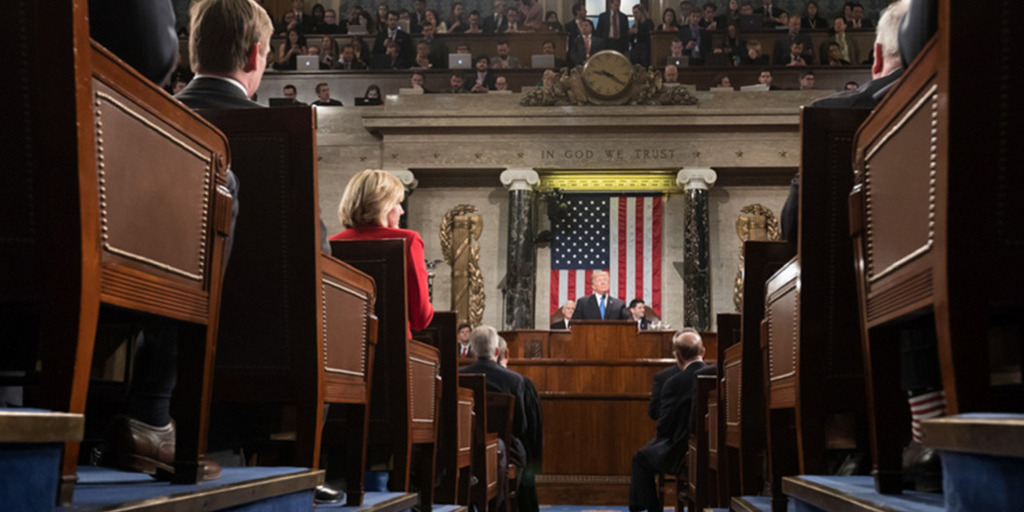 Blick in den Saal des US-Repräsentantenhauses während der Rede von US-Präsident Donald Trump zur Lage der Nation 2018. Im Vordergrund sind einige Abgeordnete von hinten zu sehen, die auf ihren Bänken sitzen und Trump zuhören. Im Hintergrund ist Donald Trump klein am Rednerpult zu erkennen.