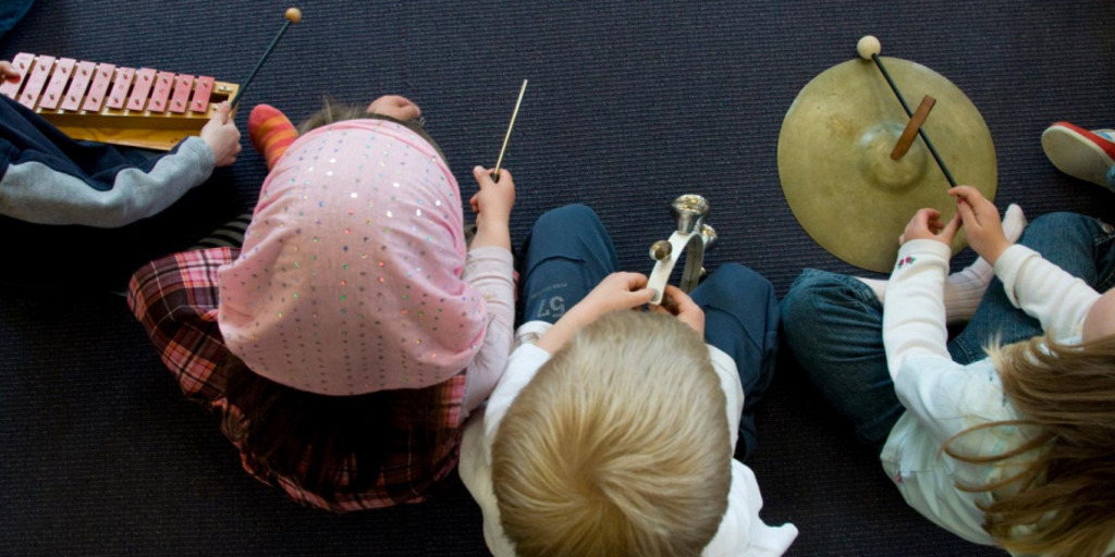 Blick von oben auf mehrere Kinder mit verschiedenen Musikinstrumenten in der Hand, einem kleinen Xylophon, einer Triangel, einem Schellenkranz mit Glocken und einer Cymbel.