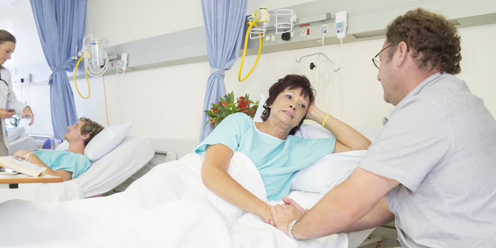 Eine Frau liegt im Patientenbett und hält die Hand ihres Mannes