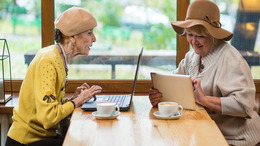 Zwei ältere Damen sitzen in einem Café an einem Tisch gegenüber und beschäftigen sich mit ihrem Laptop beziehungsweise ihrem Tablet-PC.