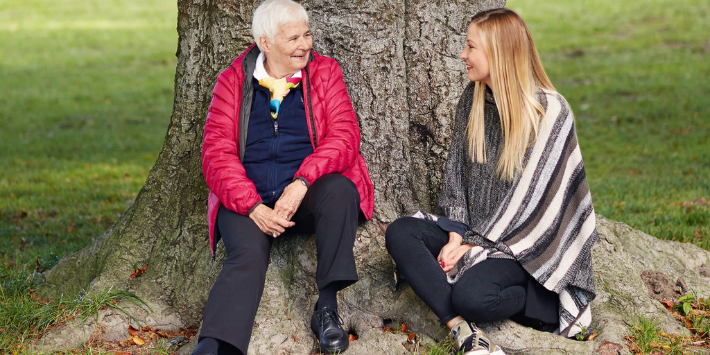 Gunda Krauss und Stephanie Neumann sitzen zu Füßen eines Baumes im Englischen Garten in München und lächeln sich an.