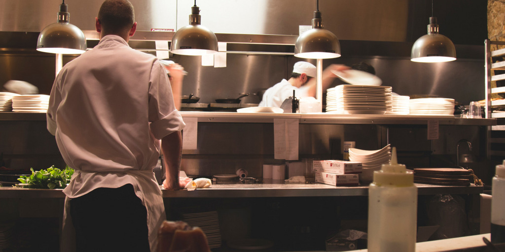Ein Koch-Azubi steht an einer Arbeitsplatte in einer Restaurant-Küche und bereitet ein Gericht zu. Er ist nur von hinten zu sehen. Im Hintergrund stehen vier Kollegen von ihm und bereiten ihre Gerichte zu, einer der Kollegen greift nach einem Teller.