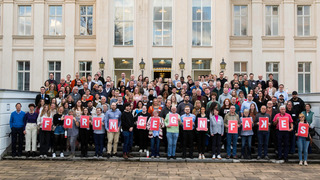 Die Teilnehmer:innen des Bürgerrates zum "Forum gegen Fakes" haben sich für ein Gruppenfoto auf den Stufen vor dem Kronprinzenpalais in Berlin aufgestellt. Einige von ihnen halten Buchstabentafeln in der Hand, die den Schriftzug "Forum gegen Fakes" ergeben.