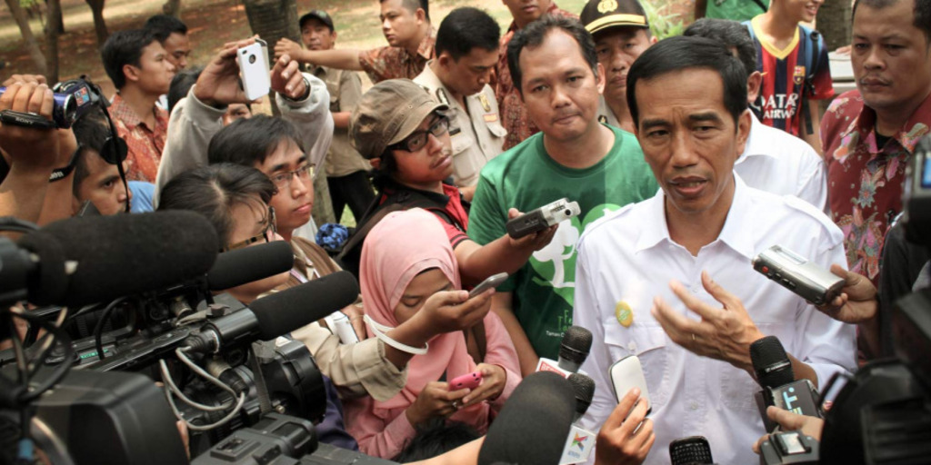 Indonesia's new president Jokowi gives an interview, surrounded by staffers, onlookers and journalists with cameras and microphones.
