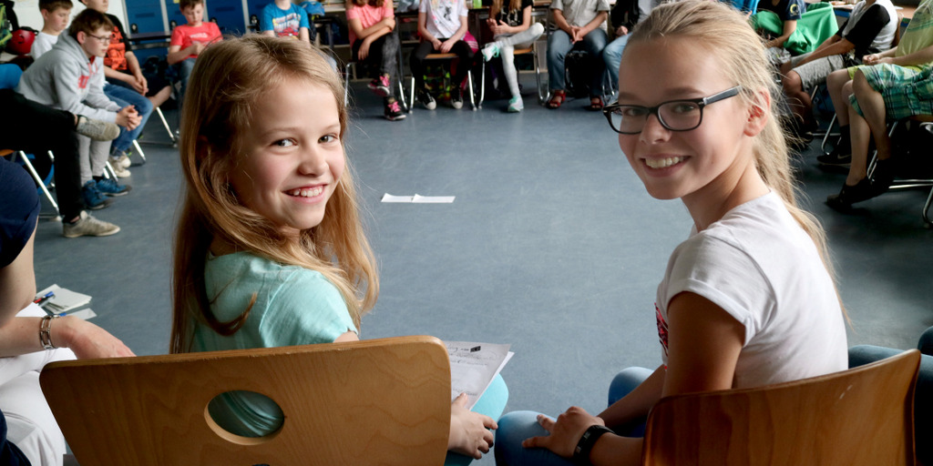 Schüler sitzen im Stuhlkreis in einem Schulzimmer. Zwei Schülerinnen haben sich in Richtung der Kamera umgedreht und lächeln.