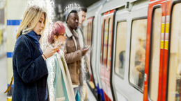 Mehrere junge Menschen stehen am U-Bahn-Gleis und schauen aufs Smartphone während ein Zug einfährt. Die Menschen werden umrahmt von Einsen und Nullen, die symbolisch für die digitale Welt stehen.