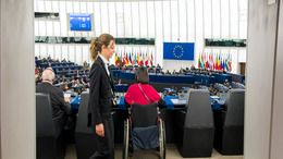 Blick von einem Gang in den Plenarsaal des EU-Parlaments in Straßbourg. Das Parlament ist voll besetzt; im Bildvordergrund sitzen zwei Parlamentarier und eine Parlamentsmitarbeiterin läuft durchs Bild.