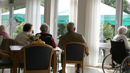 In einem Pflegeheim sitzen einige Seniorinnen und Senioren in einem Aufenthaltsraum und blicken hinaus in den sonnenbeschienenen Park.