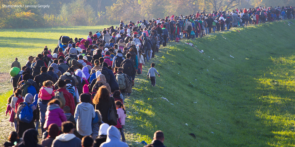 Dutzende Menschen, vermutlich Flüchtlinge, laufen in einer breiten Reihe auf einem grasbewachsenen Hügel entlang.