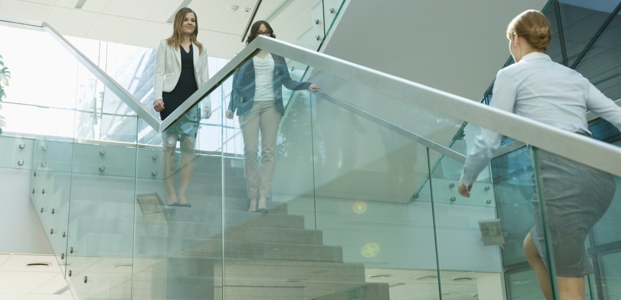 Zwei Frauen stehen auf einer Treppe. Von unten kommt ihnen auf der Treppe eine dritte Frau entgegen. Treppengeländer, Fassaden- und Dachfenster sind verglast.