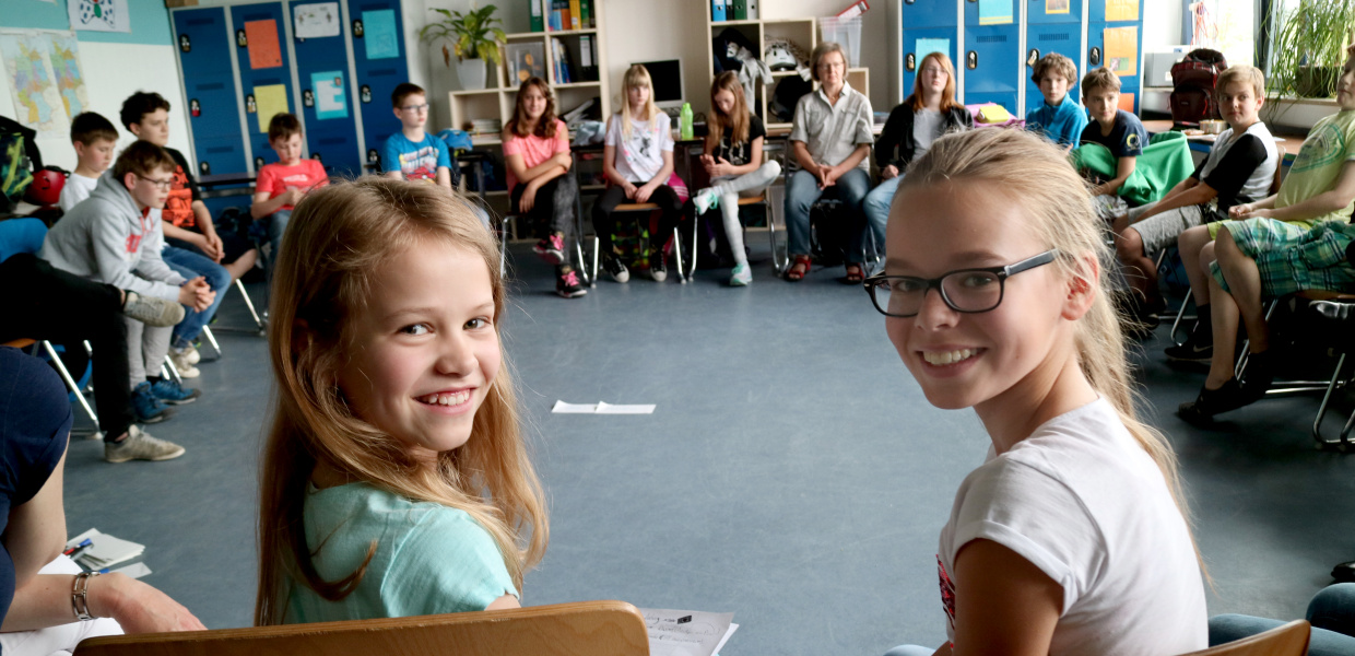 Schüler sitzen im Stuhlkreis in einem Klassenraum. Im Vordergrund sind zwei Schülerinnen zu sehen, die sich zur Kamera hin umgedreht haben und lächeln.