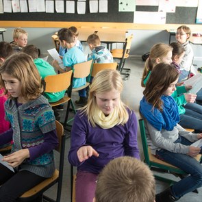 viele Kinder sitzen sich auf ihren Stühlen im Klassenraum gegenüber, sie halten ein Blatt Papier in der Hand