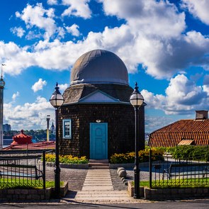 Blick auf ein Gebäude mit runder Kuppel, eingearjmt von zwei hohen Laternen. Im Hintergrund viele Wolken und die Skyline von Stockholm