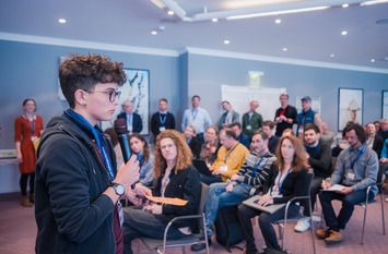 Eine Person spricht in einem Saal vor Menschen, die in Reihen sitzen