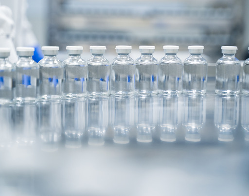 Eine Reihe kleiner, transparenter Glasfläschchen mit einer klaren Flüssigkeit und weißen Kappen steht auf einer spiegelnden Oberfläche in einem Labor oder einer Produktionsstätte. Im unscharfen Hintergrund sind vage Konturen einer Person in Schutzkleidung zu erkennen.