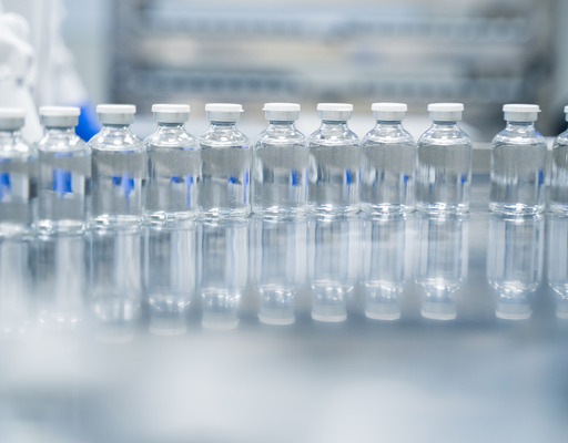 Eine Reihe kleiner, transparenter Glasfläschchen mit einer klaren Flüssigkeit und weißen Kappen steht auf einer spiegelnden Oberfläche in einem Labor oder einer Produktionsstätte. Im unscharfen Hintergrund sind vage Konturen einer Person in Schutzkleidung zu erkennen.