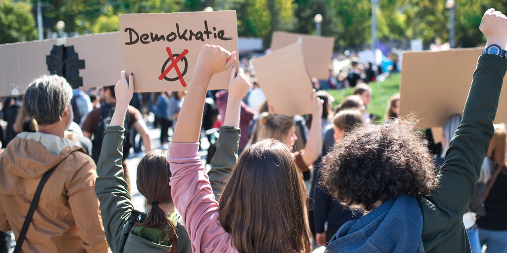 Eine Gruppe von Demonstrant:innen ist von hinten zu sehen. Sie halten Pappschilder in die Höhe, auf einem der Schilder steht das Wort "Demokratie".