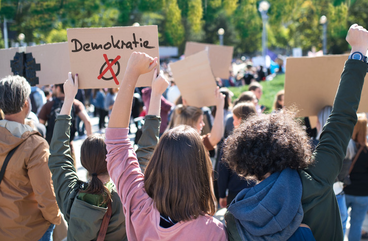 Eine Gruppe von Demonstrant:innen ist von hinten zu sehen. Sie halten Pappschilder in die H&ouml;he, auf einem der Schilder steht das Wort "Demokratie".