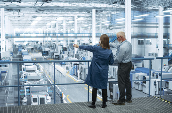 Eine Ingenieurin und ein Ingenieur stehen mit dem R&uuml;cken zur Kamera auf einem Podest und besprechen die Produktion in einer modernen, automatisierten Elektronikfertigung.