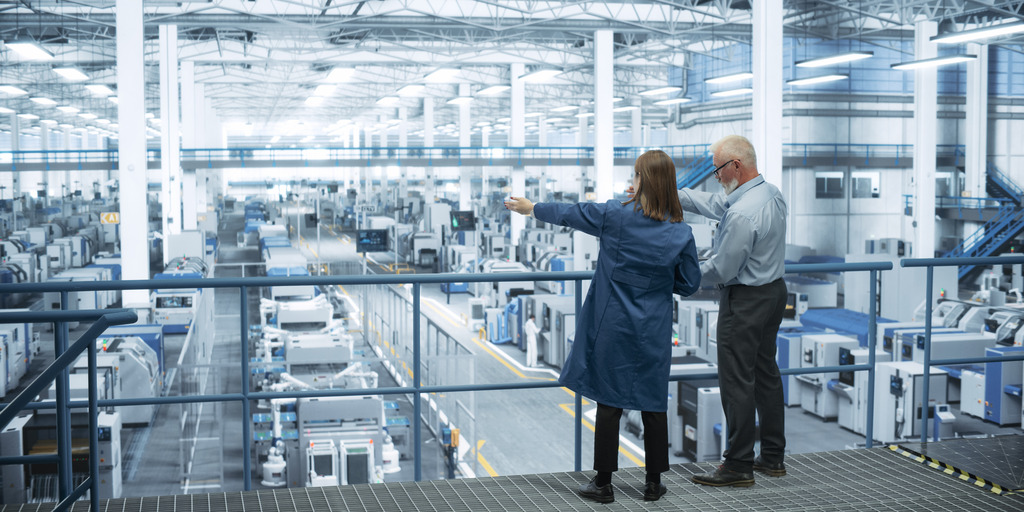 Eine Ingenieurin und ein Ingenieur stehen mit dem Rücken zur Kamera auf einem Podest und besprechen die Produktion in einer modernen, automatisierten Elektronikfertigung.