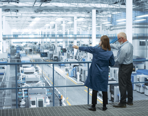 Eine Ingenieurin und ein Ingenieur stehen mit dem Rücken zur Kamera auf einem Podest und besprechen die Produktion in einer modernen, automatisierten Elektronikfertigung.