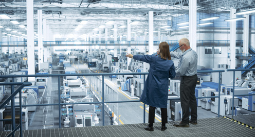 Eine Ingenieurin und ein Ingenieur stehen mit dem Rücken zur Kamera auf einem Podest und besprechen die Produktion in einer modernen, automatisierten Elektronikfertigung.