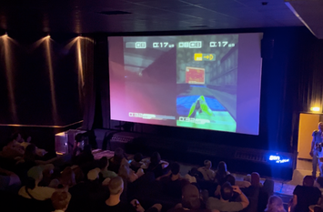 Kinosaal mit gro&szlig;er Leinwand zu sehen. Auf der Leinwand sieht man, dass zwei Personen ein Spiel gegeneinander spielen